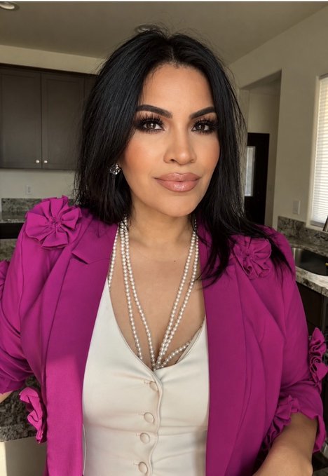 A selfie of a woman with long dark hair wearing a bright magenta blazer over a white top, accessorized with layered pearl necklaces, standing in what appears to be a kitchen.