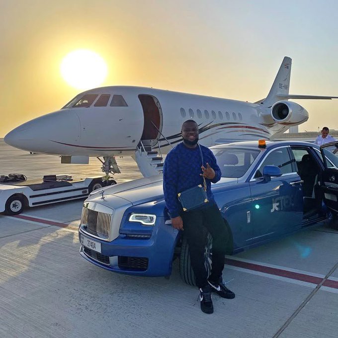 A man wearing a blue patterned sweater and black pants poses with a small blue bag in front of a blue Rolls-Royce and a private jet at sunset.