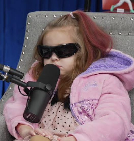 A young girl with reddish hair wearing dark sunglasses and a pink jacket sits in a gray chair in front of a podcast microphone.