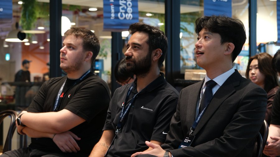 Three attendees, including representatives from Ava Labs (visible on a polo shirt) and others wearing lanyards, attentively watch a presentation at the Crypto House Seoul '26 event, aligning with the post about Team1 Türkiye's participation featuring speakers from Kite AI and Pangolin. A banner reading "Crypto House Seoul" hangs in the background, confirming the event setting.