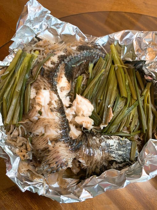 A foil-wrapped baked or grilled fish dish served over enoki mushrooms and green vegetables on a wooden table.