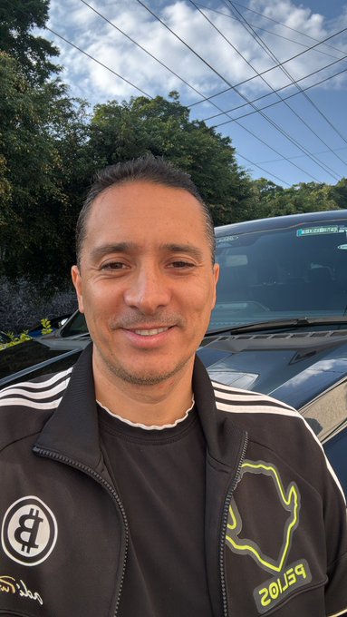 A man takes a selfie outdoors in front of a dark car, wearing a black and white zip-up jacket with green accents and the text '2015S9' visible on it.