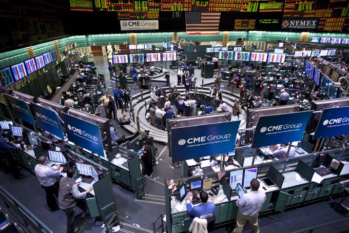 The image shows a crowded room filled with people inside of an office building, some standing by computer desks while others are walking around. There are many signs displayed throughout the area, including one that reads "BREAKING: THE WORLD'S LARGEST DERIVATIVES EXCHANGE", CME GROUP, AND ONCE. The main focus is on a large screen displaying information about Bitcoin Futures Trading and how to get started with it. It seems like an exciting event for those interested in the crypto market or financial technologies.