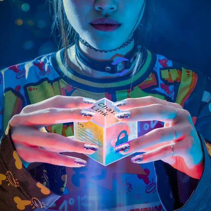 A person with glittery metallic nails holds a glowing holographic cube displaying "ChainLink" text and a padlock icon, visually symbolizing belief in $LINK cryptocurrency. The cyberpunk aesthetic with neon blue lighting reinforces the digital, futuristic nature of blockchain technology.