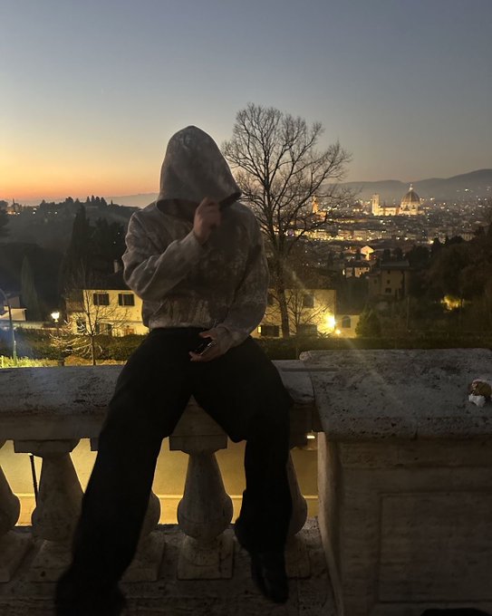 A silhouetted figure in a gray hoodie sits on a stone balustrade overlooking Florence, Italy at dusk, with the illuminated Duomo visible in the distance.