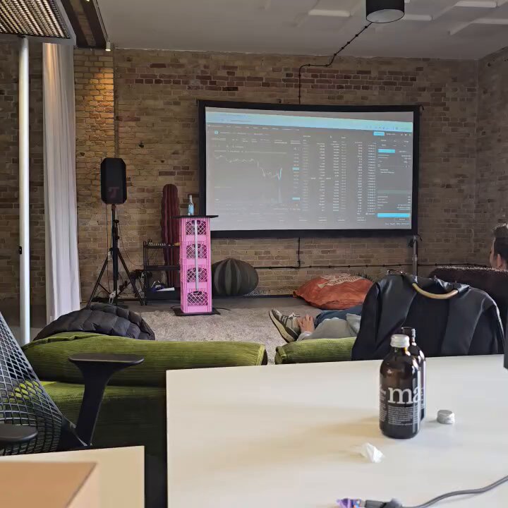 The image shows a room with a large screen displaying stock market data, likely from the cryptocurrency platform Solana. The room also features a green couch, two chairs, and a dining table. A bottle of beer can be seen placed on one of the tables. In front of the screen is a pink speaker system or sound system, which could be used for presentations or meetings in this coworking space.