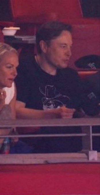 A candid photo of Elon Musk seated at a table in a dimly lit venue with red seating, wearing a dark t-shirt, appearing to be at a sporting event or show.