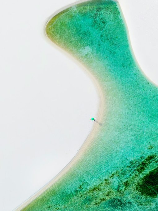 An aerial shot shows a person carrying a green umbrella walking along a white sandy beach that meets crystal-clear turquoise water.