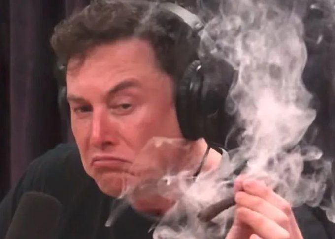 A close-up photo of Elon Musk wearing headphones and smoking a cigar during what appears to be a podcast recording, with smoke billowing around his face.