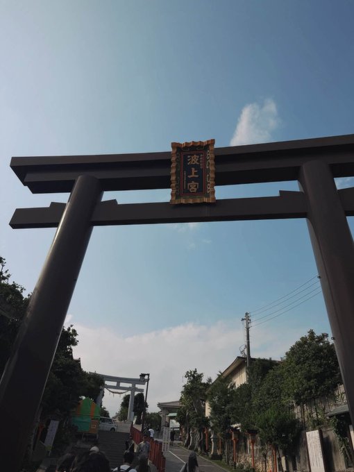 A Japanese torii gate with a plaque reading '波上宮' (Naminoue Shrine) under a blue sky, with another gate and people visible in the background along a street.