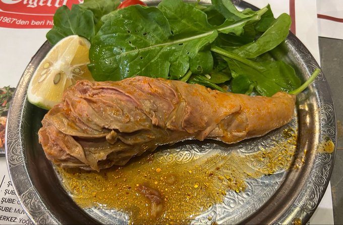 A close-up photo of a plate of food featuring a piece of roasted meat in a savory broth, garnished with fresh green arugula and a slice of lemon on a decorative metal plate.