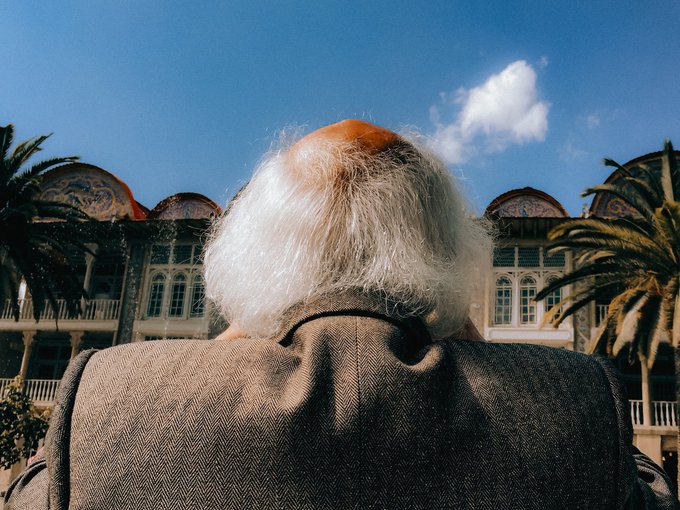 A person with voluminous white hair is photographed from behind, wearing a tweed jacket, with a historic building and palm trees in the background.