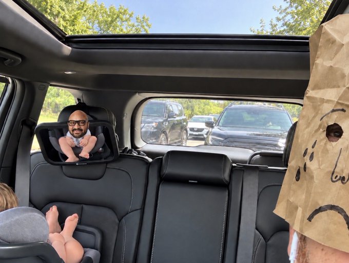 The image shows a person wearing a sad brown paper bag over their head while sitting in the back of a car, with a tablet displaying a baby's face mounted on the seat in front of them and a baby visible in a car seat. The image humorously relates to the caption about "$SUI" becoming the main character, with the person hiding their face in embarrassment or anticipation of the crypto's rise.
