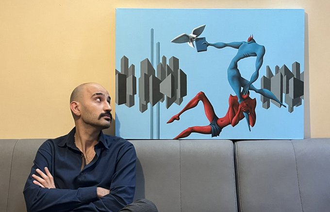 The image features a man sitting on a couch, looking at an abstract painting hanging above him. The painting depicts two people - one person wearing red and blue colors, while the other wears white and black colors. The man appears to be contemplating the artwork or simply enjoying its visual appeal. The post that accompanies this image reads: "GM Artist & Art". This suggests that the artist who created the painting is a GM artist, and the art depicted in the painting might have been inspired by GM-themed elements.