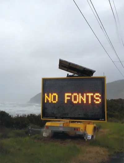 A portable electronic road sign by a coastal highway displays the message "NO FONTS" in amber LED dots, with overcast skies and the ocean in the background.