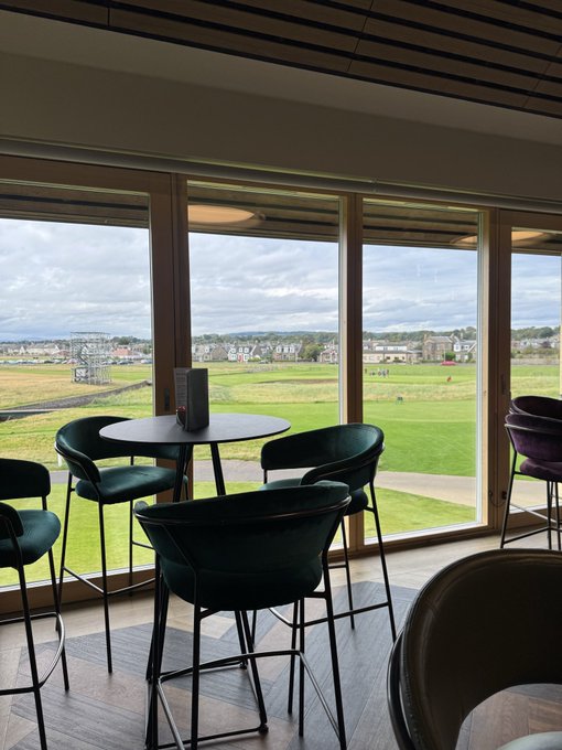 An interior view of a modern lounge or clubhouse with dark green velvet bar stools and a high table positioned in front of large floor-to-ceiling windows overlooking a golf course and town in the distance under a cloudy sky.