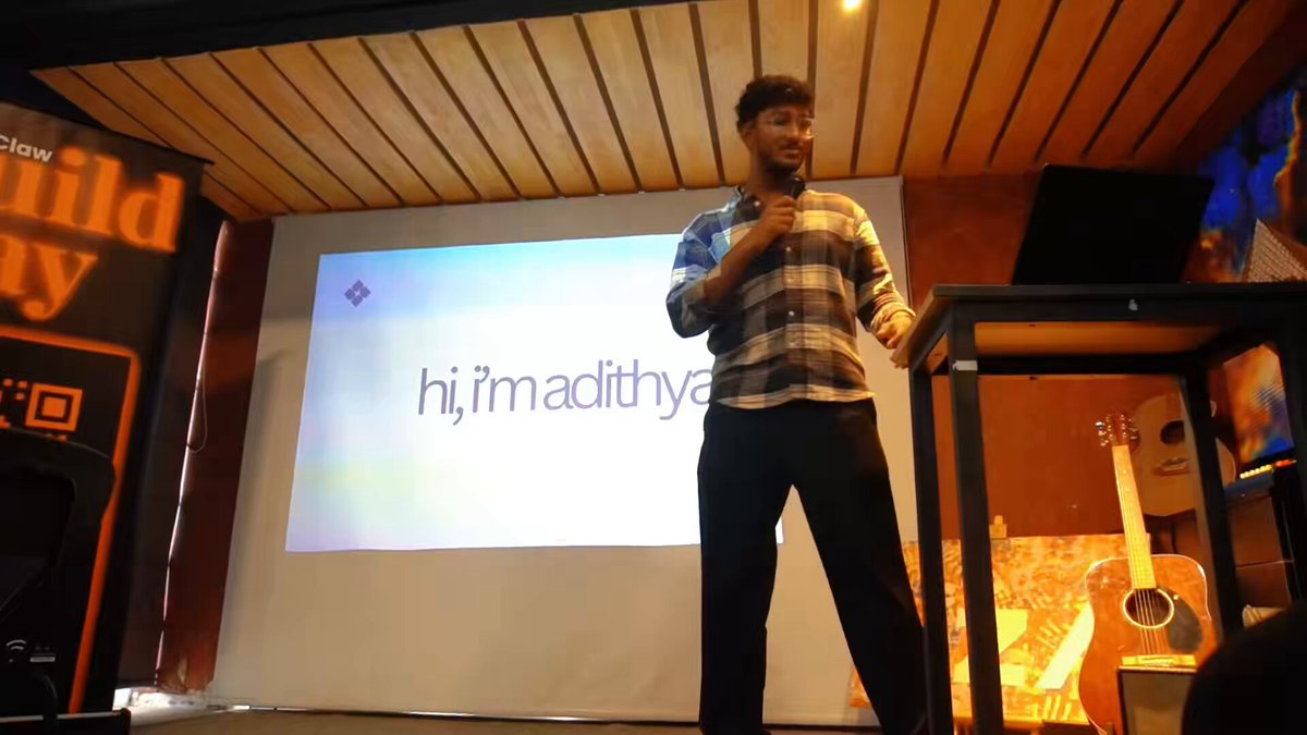 The image shows a man standing at a podium, giving a speech or presentation. He appears to be wearing a plaid shirt and black pants. The background features a large screen displaying text related to the event, such as "hi.imadithya" and "agents agents everywhere". There is also an image of a guitar on the right side of the frame.