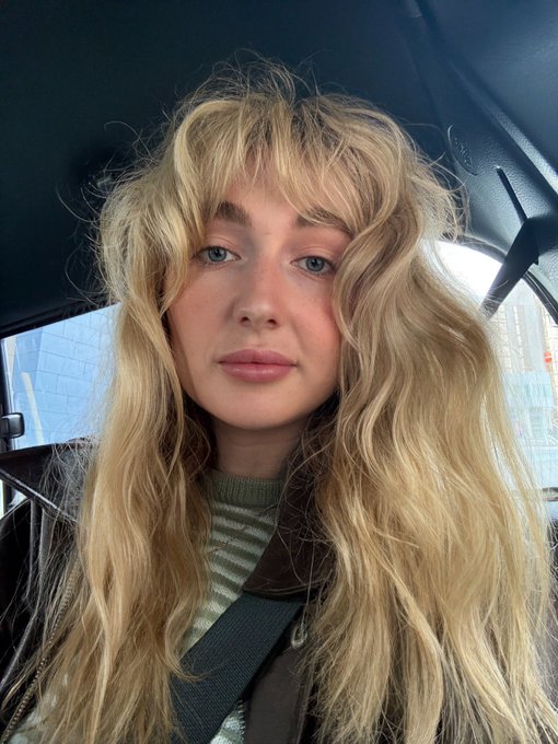 A woman with long, wavy blonde hair and curtain bangs sits in a car wearing a striped shirt under a brown leather jacket, looking straight into the camera with a neutral expression that mirrors the dry, deadpan tone of her July recap.