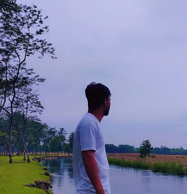 A silhouette-style photo of a man with a beard wearing a white t-shirt, standing in profile and looking out over a body of water with trees and a grassy shoreline in the background during dusk or early evening.
