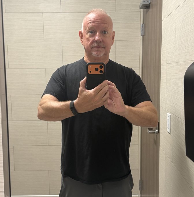 A man in a black t-shirt takes a mirror selfie with an orange-cased phone, standing in what appears to be a modern tiled restroom. The caption "Skinnier me" suggests the photo documents his fitness or weight loss progress.