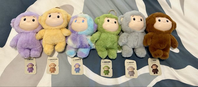 A collection of six plush "The Monke" toys (Dream, Solution, Source, Builder, Giant, and Peak) from the "Return to Origin" series is displayed in a row on a striped bed, each with an attached tag showing their name and miniature character image. The image showcases the full set highlighted in the post, with all "Source" variants noted for their unique differences.