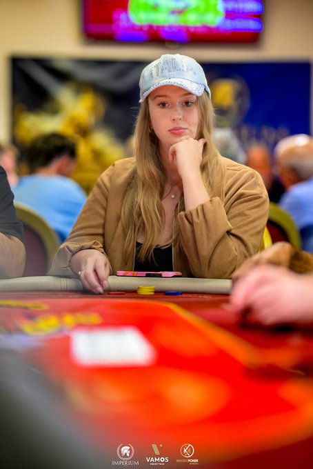 A poker player wearing a light blue cap and brown jacket sits thoughtfully at a gaming table, resting her chin on her hand while contemplating her next move, with "IMPERIUM," "VAMOS," and "POKER" logos visible at the bottom of the image, perfectly capturing the essence of "poker girl life."