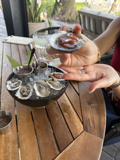 A person holds up a freshly shucked oyster topped with cocktail sauce over a bowl of oysters on ice at an outdoor table, savoring the luxury of a high-end meal that reflects the post's message about enjoying the finer things in life if you hold $ANSEM to billions.