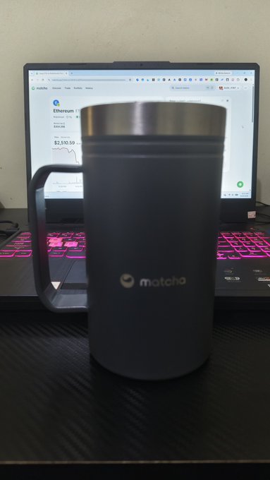 A photo of a dark gray insulated travel mug with a handle branded "matcho" sitting on a laptop keyboard with pink backlighting, with an Ethereum (ETH) price page displayed on the screen showing $2,530.59.