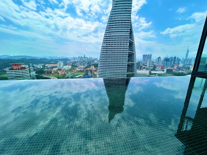A high-rise infinity pool reflecting a blue sky and clouds, with a distinctive tapered skyscraper rising from a sprawling cityscape below.