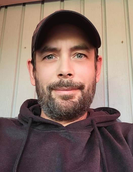 A bearded man in a maroon cap and matching hoodie takes a selfie against a light green wooden siding, his slightly graying beard and neutral expression humorously illustrating the post's joke about the stress of holding $FLR (Flare) while waiting for financial freedom.