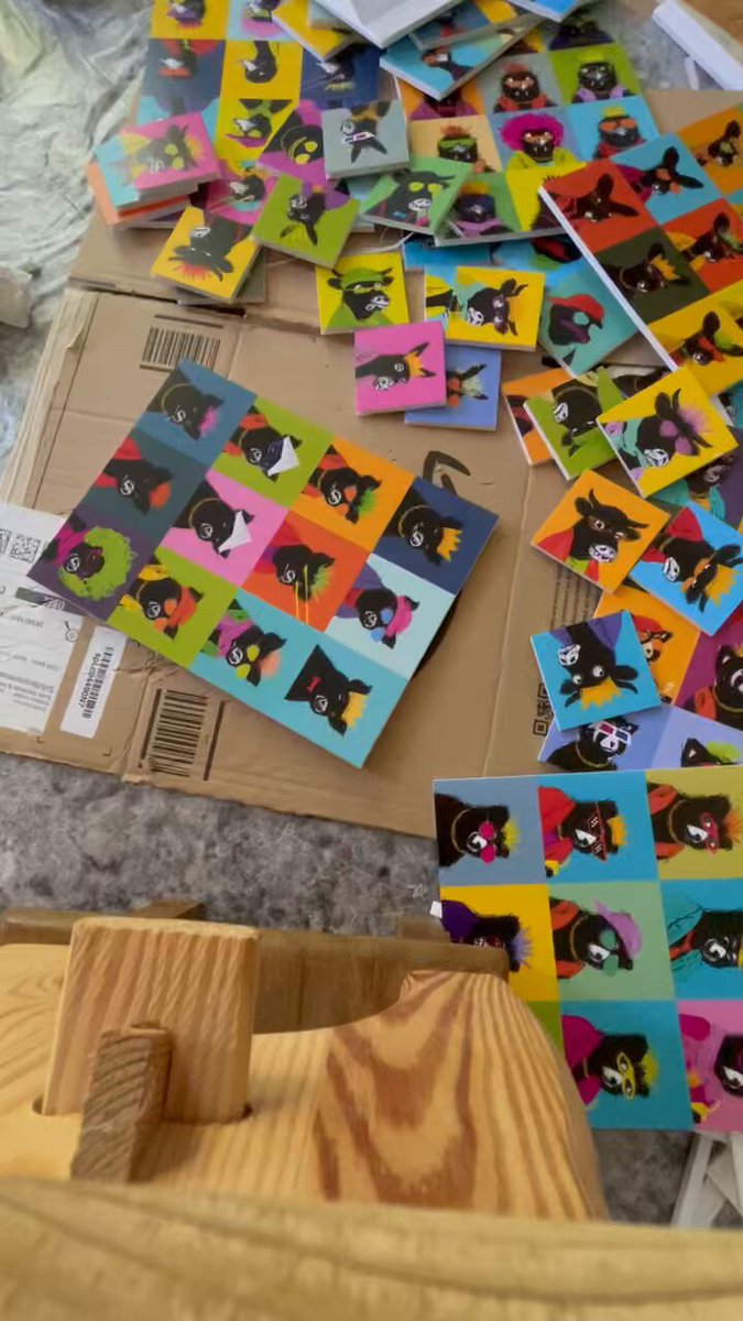 The image shows a wooden table with various animal stickers scattered across its surface, including a black cat sticker. Some of these stickers are overlapping each other, creating an interesting visual effect. The table appears to be covered by cardboard boxes or a similar material, which is also present in the scene.