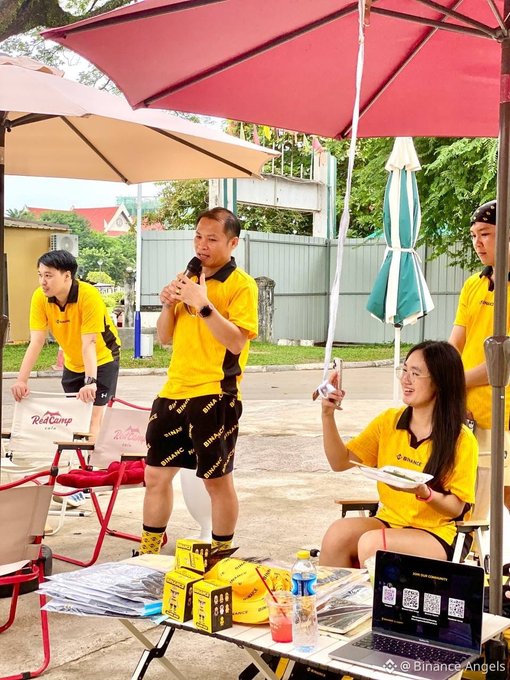 Members wearing yellow Binance shirts gather under red and white umbrellas for a community event, with a man speaking into a microphone and a smiling woman holding a phone, while boxes of Binance-branded items and a laptop displaying QR codes sit on the table in front of them, capturing the lively "Run. Connect. Have fun." spirit of the Laos Angels City Run.
