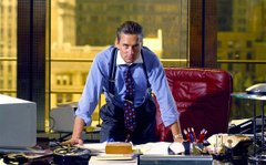 A man in a blue shirt and suspenders sits at a cluttered office desk, looking toward the camera with papers and office supplies scattered around him.