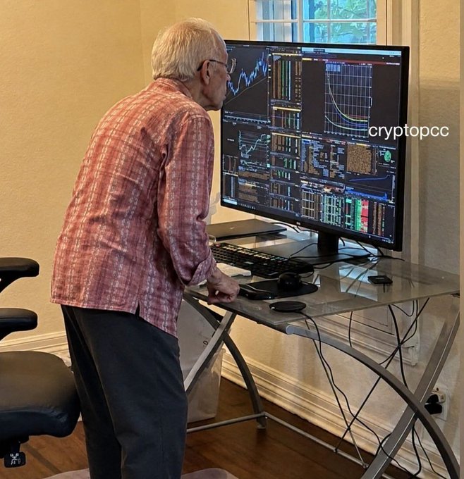 An older man in a plaid shirt standing at a desk reviewing cryptocurrency trading charts on a large monitor, with the text 'cryptopcc' visible.
