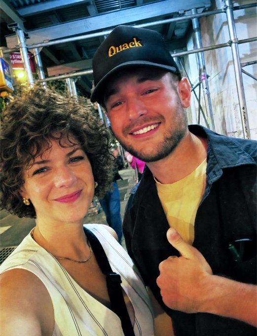 A smiling woman with curly hair takes a selfie with a smiling man wearing a black cap that says 'Quack' on it; they are standing on a city street at night.