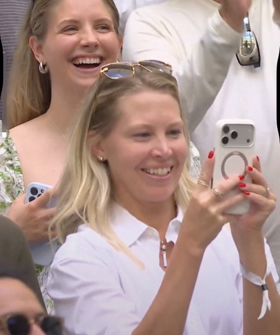 Two blonde women smile and laugh while holding smartphones, with the woman in front sporting red nail polish and a white shirt.