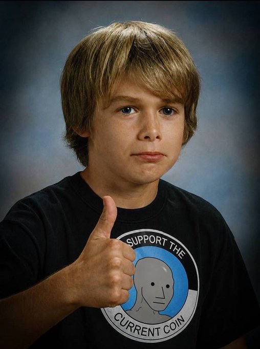 A young boy with blonde hair giving a thumbs up while wearing a black t-shirt that reads 'SUPPORT THE CURRENT COIN'.