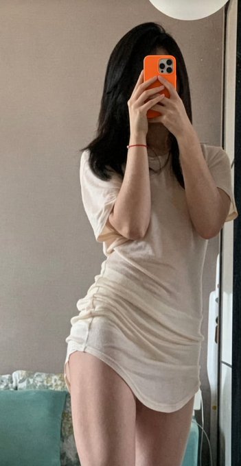 A person with long dark hair takes a mirror selfie wearing an oversized beige t-shirt that falls off one shoulder, holding an orange iPhone with a triple-camera setup to cover their face.