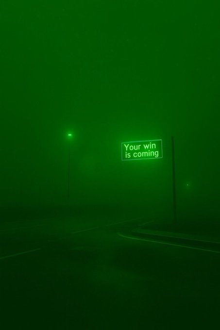 A foggy, green-tinted road scene at night features a glowing street sign reading "Your win is coming," visually echoing the post's motivational message about perseverance. The solitary streetlight and curving road reinforce the journey toward an anticipated triumph.