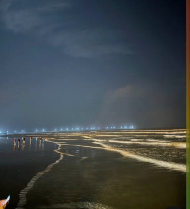 A nighttime beach scene with people walking along the shoreline, waves reflecting distant city lights under a cloudy sky.