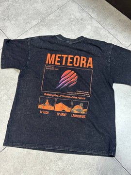 A dark grey t-shirt laid on a tiled floor featuring orange and purple graphics with the text 'METEORA', 'Building the LP Toolkit of the Future', and a city skyline labeled 'LIQUIDITY', with location markers 'LI TECH', 'LI ANALYTICS', and 'LAUNCHPAD'.