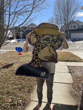A person outdoors holding up a large cartoon cutout of a monkey wearing a yellow beanie and smoking a cigarette, covering their face in a suburban neighborhood setting.