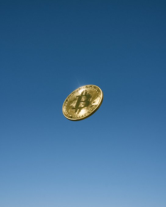 A golden Bitcoin coin floats against a clear blue sky, with the iconic "B" symbol prominently displayed on its face. The image visually represents the cryptocurrency's soaring value, evoking the idea of "to the moon."