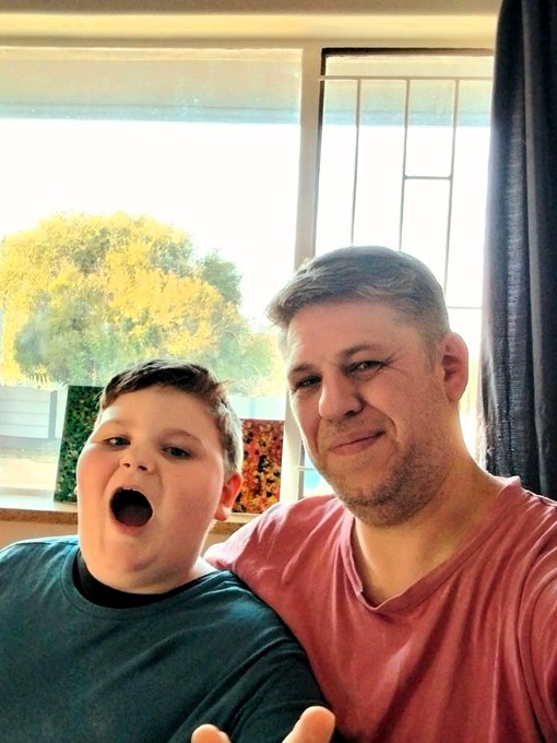 A man in a coral shirt smiles warmly alongside a young boy in a teal sweater who is making a playful, open-mouthed expression. Behind them, a window reveals sunlit trees, and colorful paintings lean on the windowsill—capturing the relaxed family creativity mentioned in the caption.