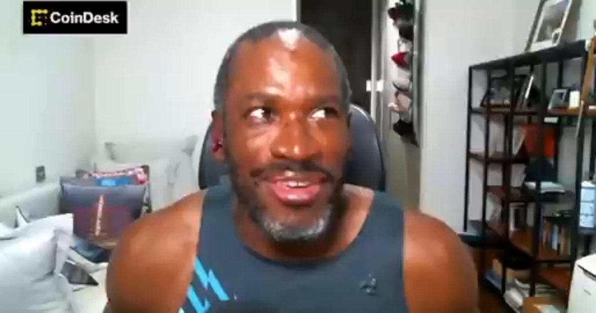 The image shows a man wearing a black tank top sitting at a desk with a computer monitor behind him. The man appears to be smiling or making a funny face as he looks directly into the camera. The background of the image features a bookshelf filled with various items, including several books and a few bottles.