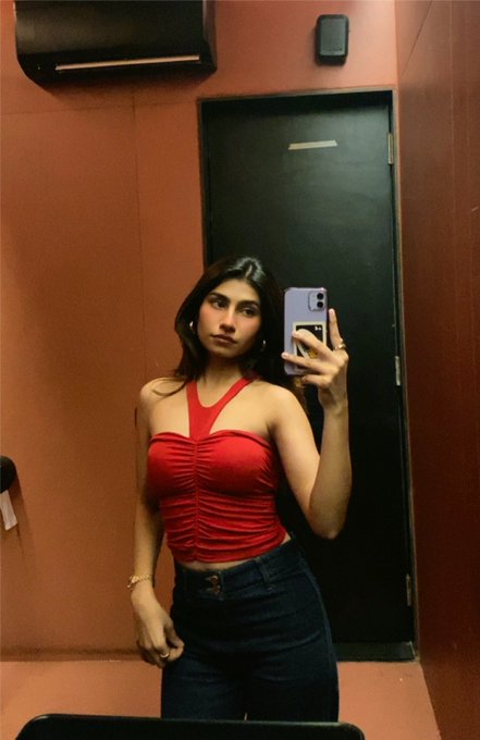 A woman takes a mirror selfie wearing a red ruched halter top and dark jeans in a fitting room, posing with her phone in a lavender case. The casual, confident vibe complements her post's playful "gm to those who gm back" message, matching the energy of a chill morning check-in with style.