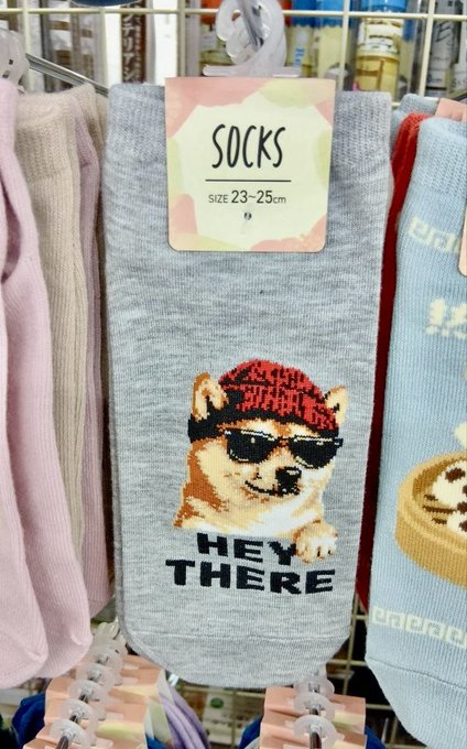 A package of grey socks hangs on a display rack; the label reads "SOCKS SIZE 23~25cm," and the socks feature a graphic of a dog wearing sunglasses and a red bandana with the text "HEY THERE."
