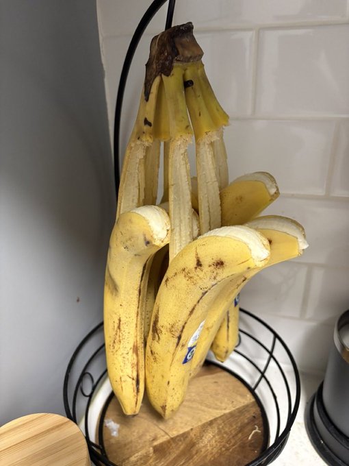 A bunch of ripening yellow bananas hanging from a black wire fruit basket in a kitchen with a white tiled backsplash.