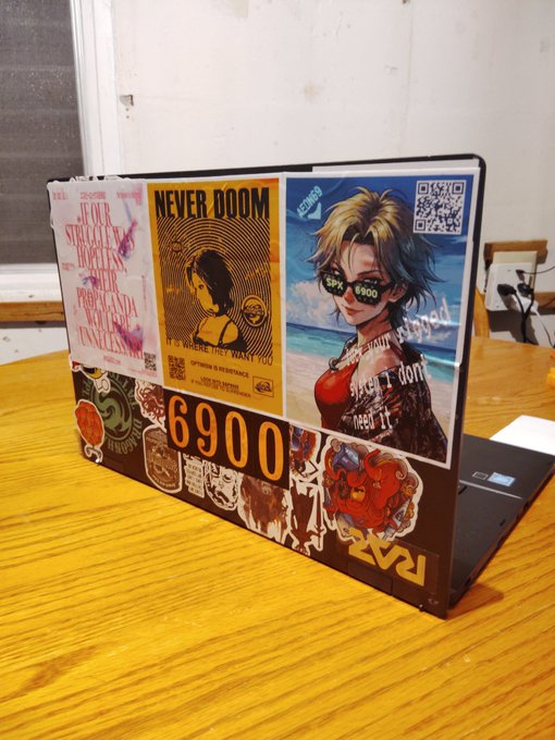 A black laptop covered in colorful anime and pop-culture stickers sits on a wooden desk, featuring prominent stickers with the number '6900' and text including 'NEVER DOOM' and 'if its where you want to'.