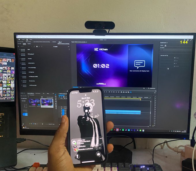 A workspace setup showing a hand holding an iPhone displaying a lock screen with the time '5:33' over a background of a large curved monitor running editing software with 'KICKCHAIN' and '01:02' visible on the screen.
